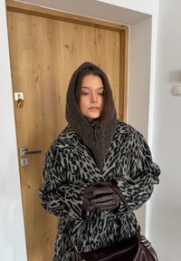 Manteau en laine imprimé léopard, capuche en tricot marron avec cordon de serrage, gants en cuir marron, tenant un sac à main marron foncé, debout à côté d'une porte en bois.