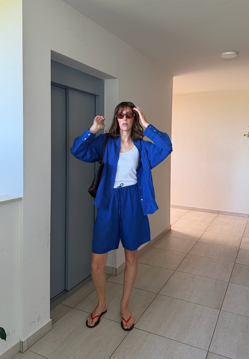 Femme portant un short bleu et une chemise, un haut blanc, des lunettes de soleil rouges et des tongs, debout près d'un ascenseur fermé dans un couloir carrelé, ajustant ses lunettes de soleil.