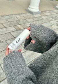 Η αντηλιακή κρέμα ILIA σε λευκό κυλινδρικό μπουκάλι κρατείται από ένα χέρι που φοράει έναν χοντρό, υφασμάτινο γκρι πουλόβερ με φαρδιές, ατελείωτες μανσέτες.