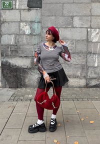 Čierno-biele pruhované tričko s priehľadnými kvetinovými rukávmi, červený baret, čierna mini sukňa, bordové pančuchy, robustné čierne platformové topánky a červená chlpatá taška.