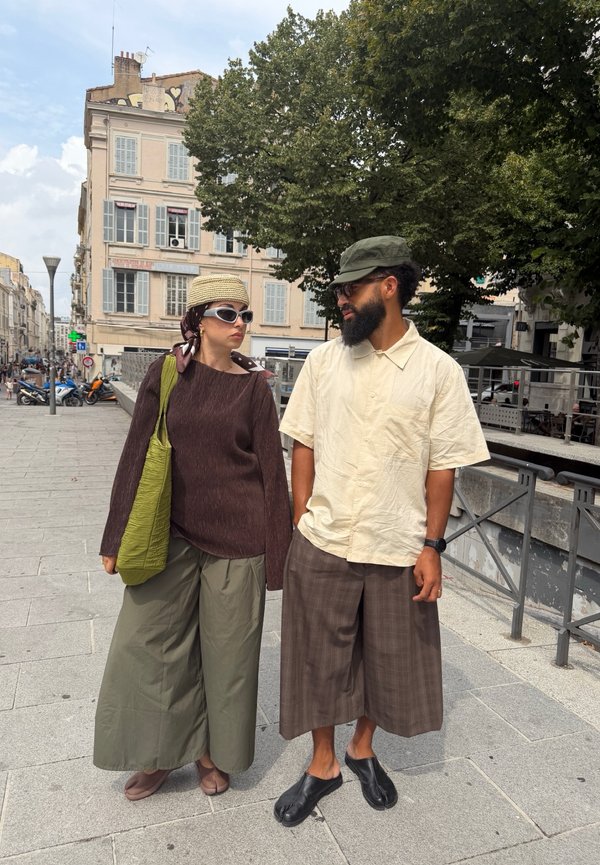 Deux personnes portant des pantalons larges et des chapeaux se tiennent sur un trottoir en ville, avec des bâtiments et des arbres en arrière-plan.