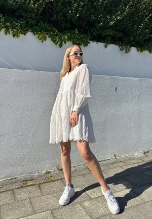Vestido de encaje blanco con manga larga, diseño en capas, dobladillo con puntilla, combinado con zapatillas blancas, frente a una pared blanca texturizada y vegetación.