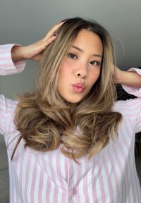 Długie, falujące włosy z blond refleksami, ubrana w bladoróżową koszulę w białe paski z guzikami, pozująca z rękami we włosach, lekko zastępione usta.