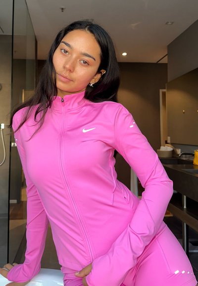 Mujer joven con una chaqueta deportiva de cierre frontal en un brillante color rosa, inclinándose hacia adelante en interiores con las manos en las caderas, cabello largo y oscuro, y expresión neutral.