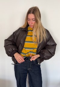 Doudoune marron à col montant, portée sur une chemise rayée jaune et verte, associée à un jean en denim foncé avec une taille à cordon de serrage.