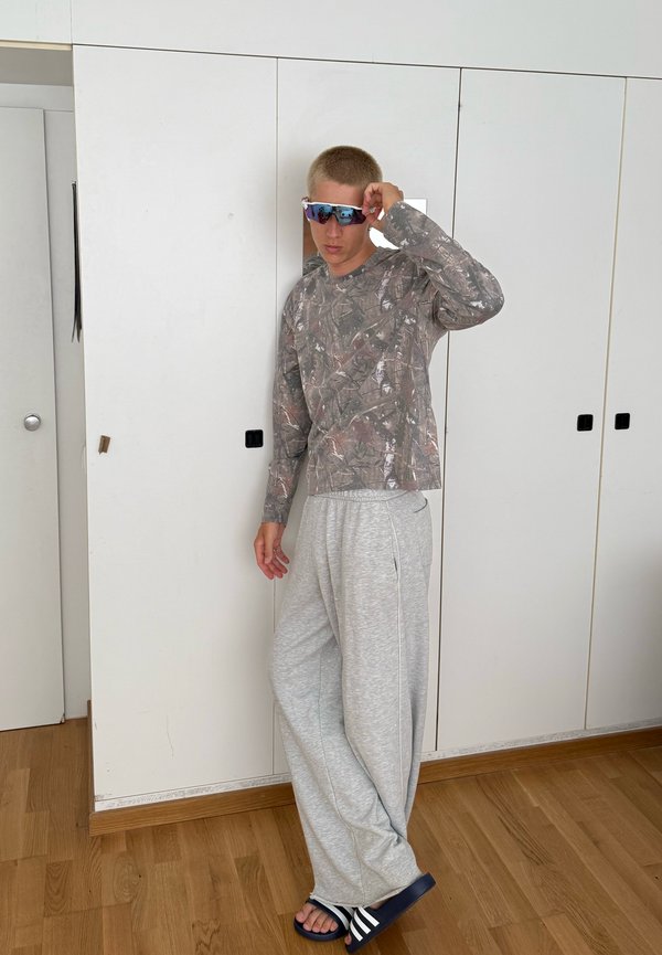 Jeune homme portant une chemise camouflage à manches longues, un pantalon de jogging gris, des claquettes bleu marine et des lunettes de soleil réfléchissantes, debout à l'intérieur à côté de placards blancs.