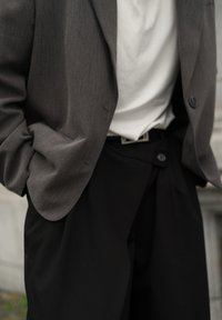 Šedé sako z texturované látky, doplněné bílým tričkem a volnými černými kalhotami, s viditelným páskem a knoflíkovými detaily.