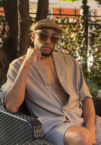 Μπεζ κοντομάνικο πουκάμισο με βαθύ V-λαιμό και λεπτομέρεια από κουμπιά, σε συνδυασμό με αντίστοιχες σορτσάκι. Το άτομο φοράει μεγάλα γυαλιά ηλίου και καπέλο σε απόχρωση ταμπά.