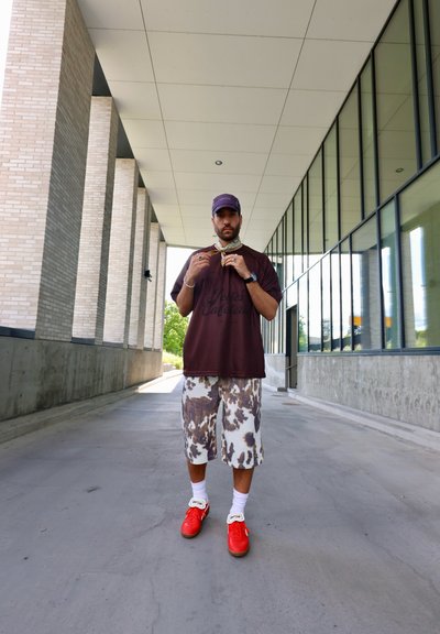 Camiseta marrón oversized con texto, pantalones cortos de camuflaje blanco, zapatillas rojas y accesorios que incluyen un collar y una gorra en un entorno urbano.