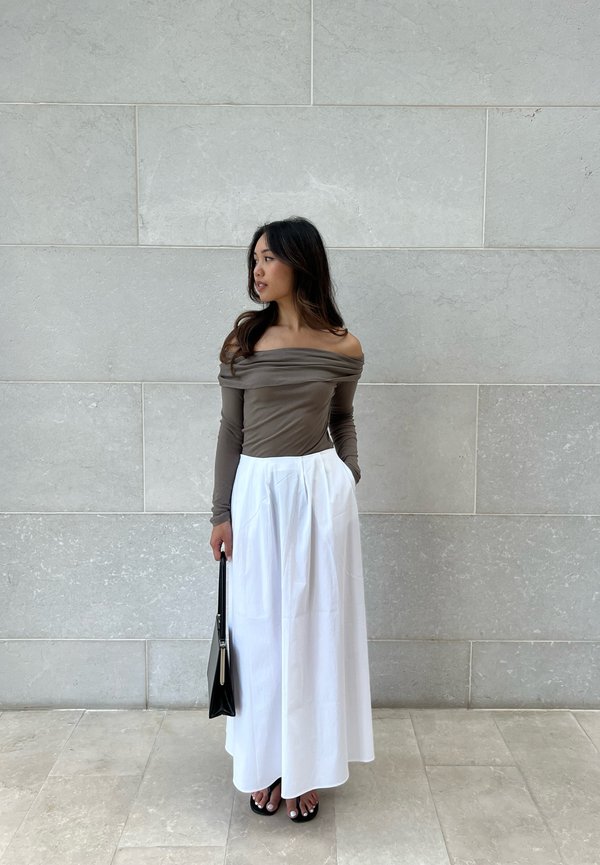 Langärmliges, schulterfreies, braunes Oberteil, kombiniert mit einem weißen, bodenlangen Rock. Eine schwarze Handtasche und offene Sandalen runden das Outfit ab.