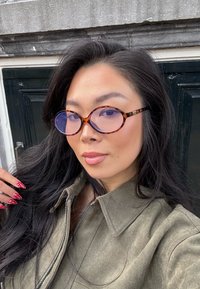 Lunettes rondes en écaille de tortue avec des verres teintés bleus, portées par une personne dans une veste olive. Cheveux longs et foncés, maquillage léger et ongles à pois.
