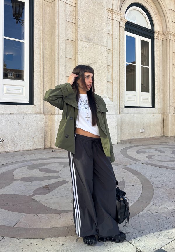 Tânără îmbrăcată într-o jachetă militară, tricou alb cu grafică, pantaloni largi negri de trening cu dungi albe, stând pe un podea din piatră modelată lângă ferestre cu arcade.