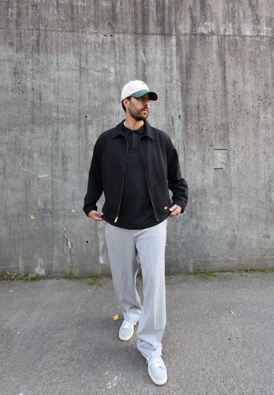 Chaqueta negra con cremallera sobre una camiseta negra, pantalones rectos gris claro y zapatillas grises, complementados con una gorra blanca con visera verde.