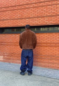 Veste en polaire marron avec col montant, associée à un jean bleu décontracté. Chaussures noires avec des accents verts. Fond en brique rouge.
