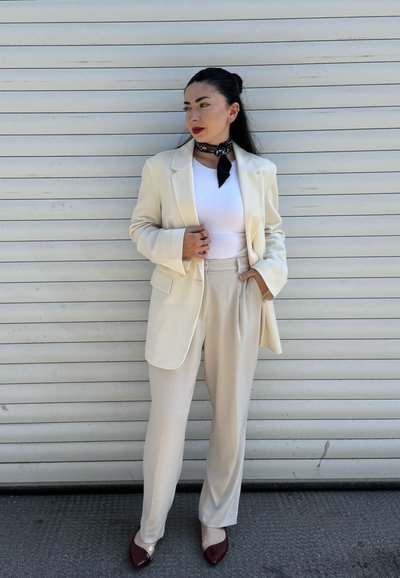 Blazer beige ajustado sobre una blusa blanca, combinado con pantalones claros y tacones burdeos puntiagudos. Un pañuelo negro acentúa el conjunto.