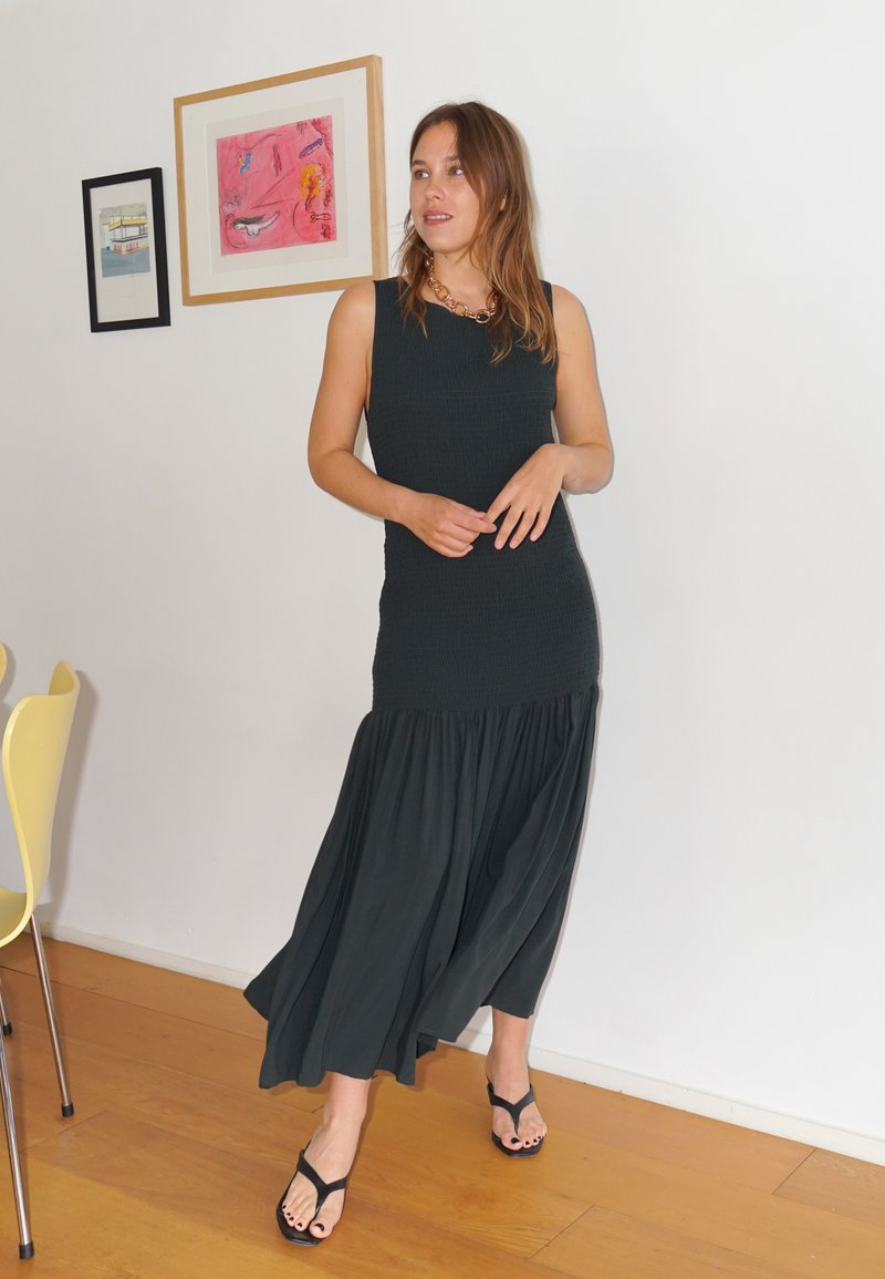 Musta maksimekko, jossa on tiukka yläosa ja valuvasta, röyhelöisestä materiaalista valmistettu hame. Yhdistetty mustiin flip-flop sandaaleihin. Taustalla neutraali seinä ja puulattia.