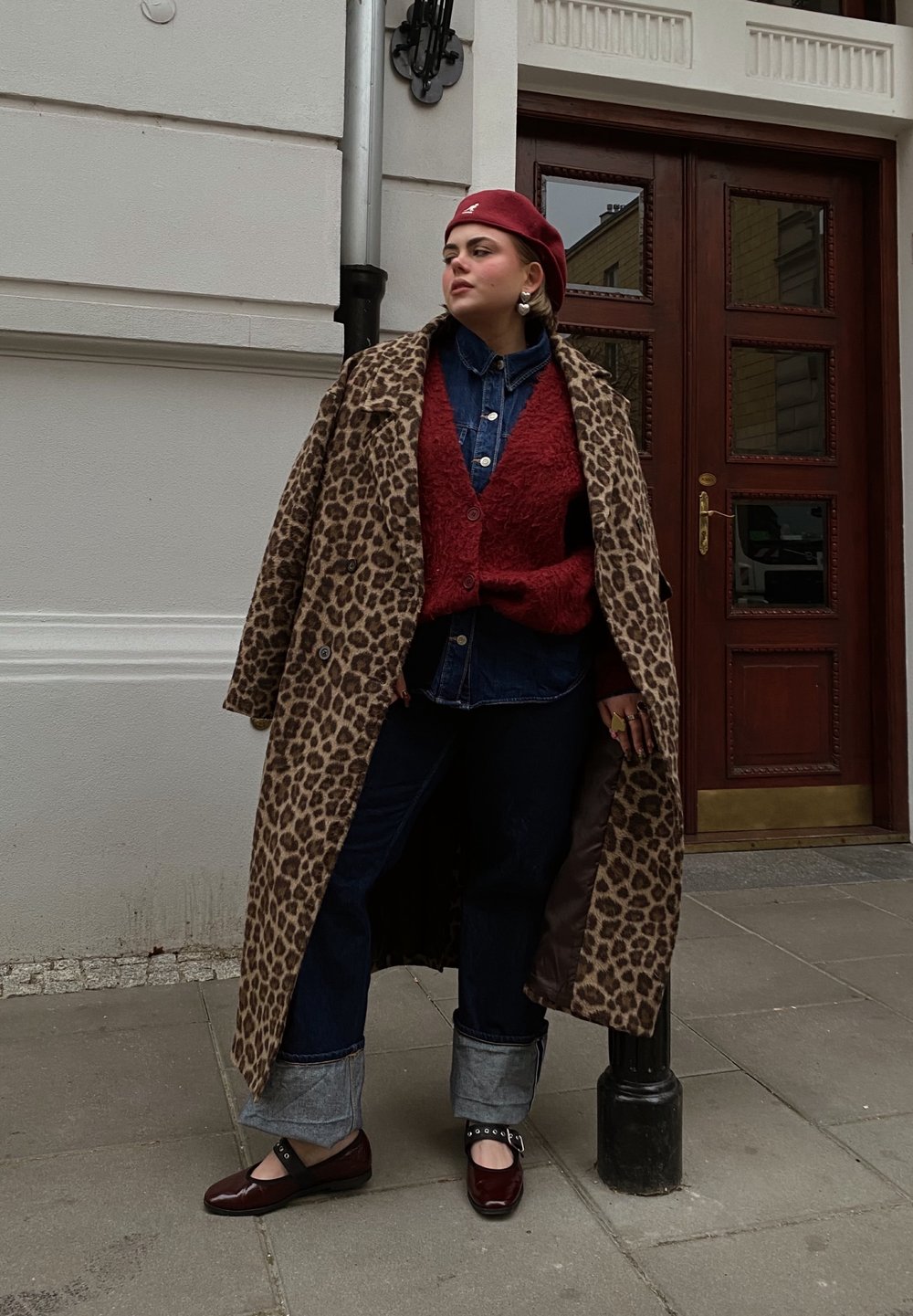 Manteau à imprimé léopard, cardigan rouge texturé, chemise en denim, jeans noirs avec ourlets à revers, et chaussures bordeaux avec boucles. Cadre urbain.