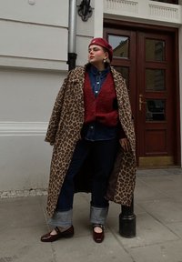 Manteau à imprimé léopard, cardigan rouge texturé, chemise en denim, jeans noirs avec ourlets à revers, et chaussures bordeaux avec boucles. Cadre urbain.