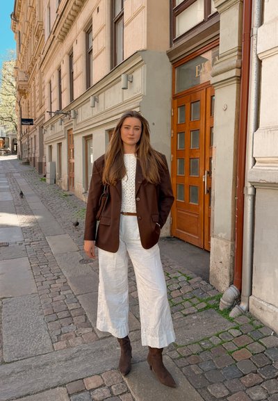 Joven con el pelo largo lleva una blazer marrón, top de punto blanco, pantalones blancos anchos y botines marrones, de pie en una acera adoquinada.