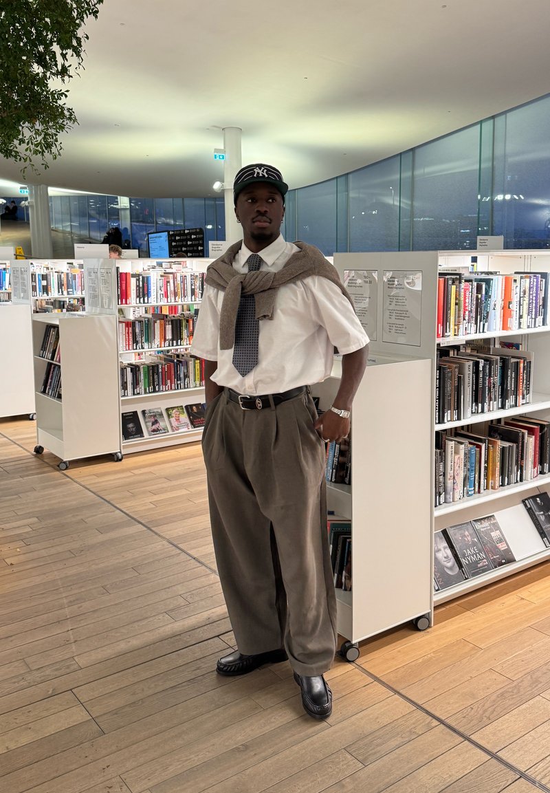 Junger Mann mit Cap, Krawatte und Pullover über den Schultern steht in einer modernen Bibliothek, umgeben von Bücherregalen und einem Holzboden.
