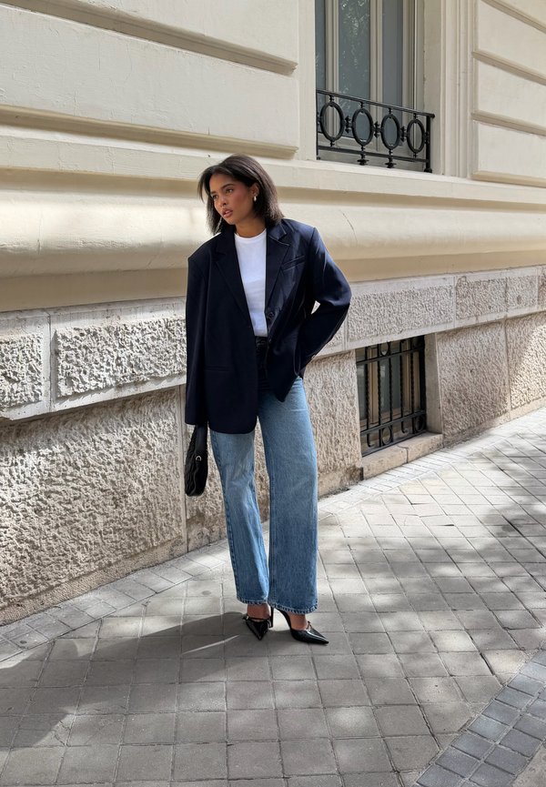 Una mujer con una americana azul marino, un top blanco, unos vaqueros de pernera ancha y zapatos de tacón puntiagudos posa junto a un edificio de piedra mientras sostiene un bolso negro.
