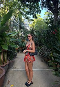 Femme en lunettes de soleil et robe colorée debout sur un chemin de jardin entouré de plantes vertes luxuriantes en pots sous un ciel clair.
