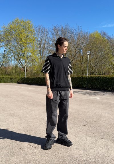 Persona joven con tatuajes que lleva un chaleco negro sobre una camisa de rayas y pantalones negros, de pie en un aparcamiento vacío bañado por el sol con árboles al fondo.