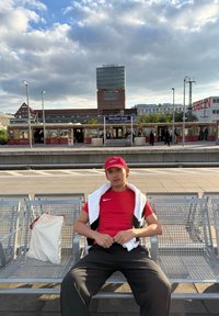 Giovane uomo con maglietta rossa e cappellino seduto su una panchina di metallo alla stazione ferroviaria di Warschauer Straße con una borsa bianca accanto a lui.