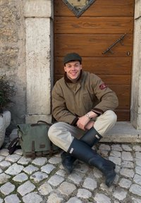 Jeune homme en veste beige et bottes de pluie noires assis en tailleur sur un pavé en pierre, souriant devant une porte en bois avec un sac vert à côté de lui.