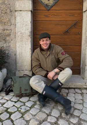 Joven con chaqueta beige y botas de lluvia negras sentado con las piernas cruzadas sobre un pavimento de piedra, sonriendo frente a una puerta de madera con una bolsa verde a su lado.