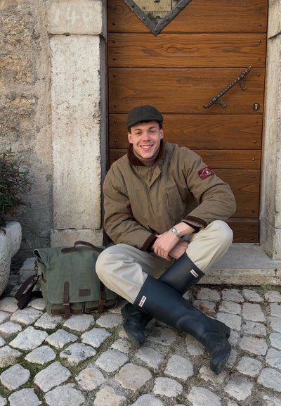 Joven con chaqueta beige y botas de lluvia negras sentado con las piernas cruzadas sobre un pavimento de piedra, sonriendo frente a una puerta de madera con una bolsa verde a su lado.