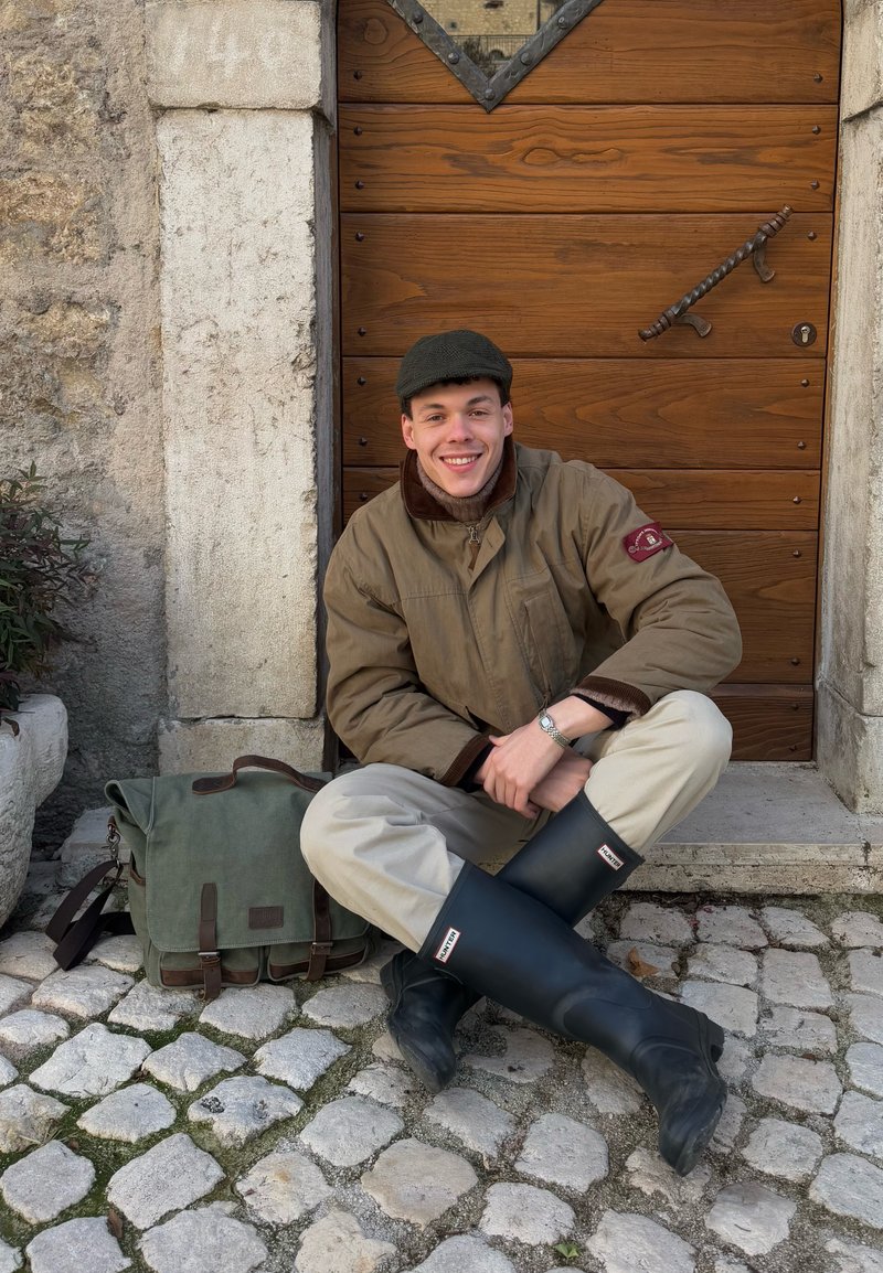 Jeune homme en veste beige et bottes de pluie noires assis en tailleur sur un pavé en pierre, souriant devant une porte en bois avec un sac vert à côté de lui.