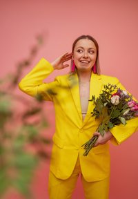 Abito giallo con giacca aderente e pantaloni coordinati. Tiene un mazzo di rose e verde. Sullo sfondo si vede un muro rosa.