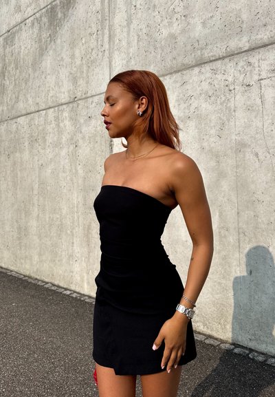 Vestido negro sin tirantes con un corsé ajustado y detalle fruncido. Combinado con un reloj plateado y pendientes de perno. Al fondo se puede ver una pared texturizada de color gris.