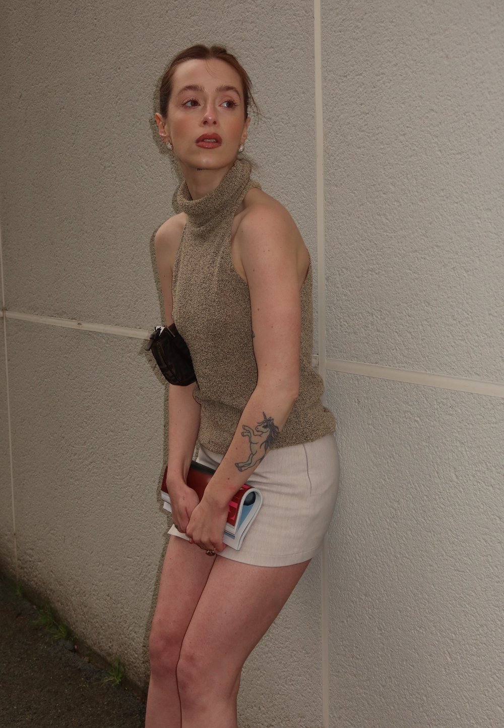 Beige ilmanhihaista pooloneuletta, vaaleaa minihametta, pitämässä lehtiä, pieni musta laukku ja näkyvä tatuointi vasemmassa kädessä.