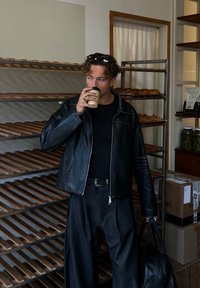 Svart skinnjakke over en svart skjorte, sammen med løse svarte bukser. Holder en kaffekopp, står i et bakeri med trehyller.