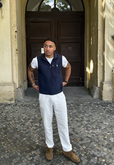 Chaleco de marino con detalles de cremallera y bolsillo, camiseta blanca, pantalones de lino blancos y zapatos de ante marrones, de pie en un patio de piedra texturizada.