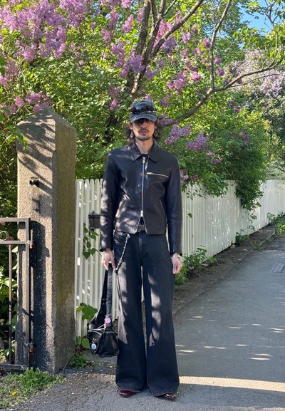Chaqueta de cuero negra con cremallera y herrajes plateados, combinada con pantalones oscuros de pierna ancha. Sosteniendo un pequeño bolso negro. Rodeada de flores moradas.