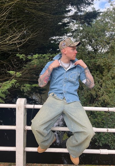 Camisa de mezclilla azul claro con cierre de botones, pantalones anchos azul claro y una gorra de camuflaje. Se pueden ver cadenas y tatuajes.