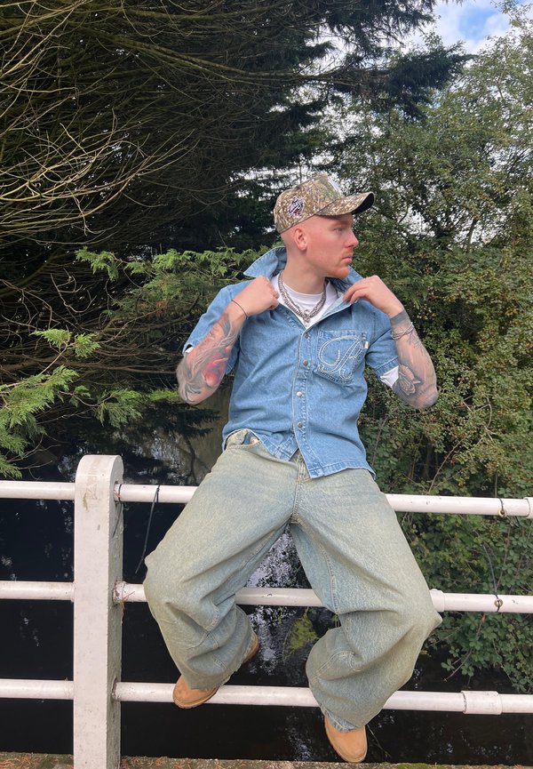 Camicia di jeans azzurro chiaro con chiusura a bottoni, pantaloni oversize azzurro chiaro e berretto in camouflage. Catene e tatuaggi sono visibili.