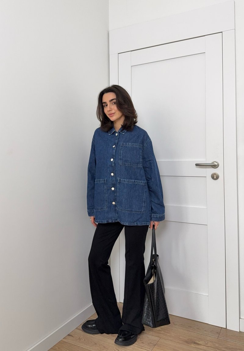 Übergroßes denim Hemd in Dunkelblau mit Knopfverschluss und zwei vorderen Taschen, kombiniert mit schwarzen Hosen und einer schwarzen Tragetasche. Flache Schuhe sind ebenfalls enthalten.