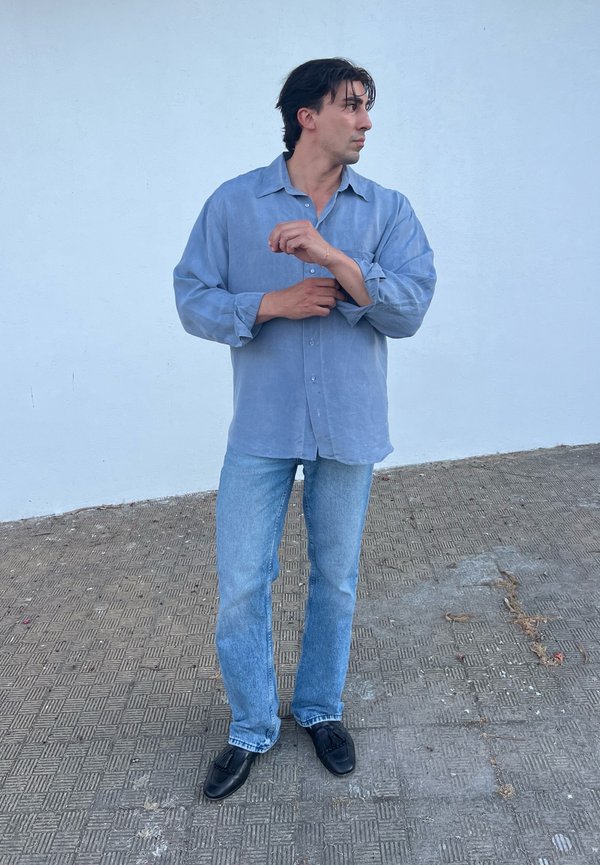 Mann steht auf gefliestem Boden, krempelt das Ärmel eines hellblauen Hemdes hoch, trägt Jeans und schwarze Schuhe, schaut nach rechts.