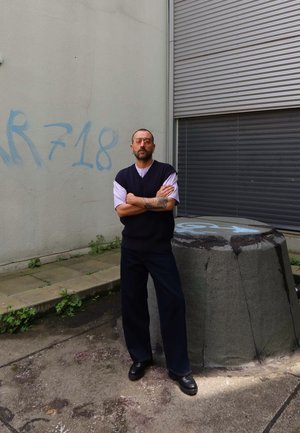 Persona con un chaleco azul marino sobre una camisa blanca, pantalones negros y zapatos, de pie junto a un bloque de concreto texturizado contra una pared gris.