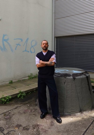 Persona con un chaleco azul marino sobre una camisa blanca, pantalones negros y zapatos, de pie junto a un bloque de concreto texturizado contra una pared gris.