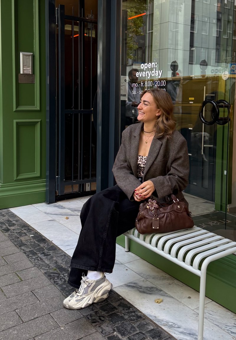 Brun uld blazer, leopardprintet top, sorte vidde bukser, hvide sokker, grå sneakers. Brun taske med blomsterdetaljer på en metalbænk.