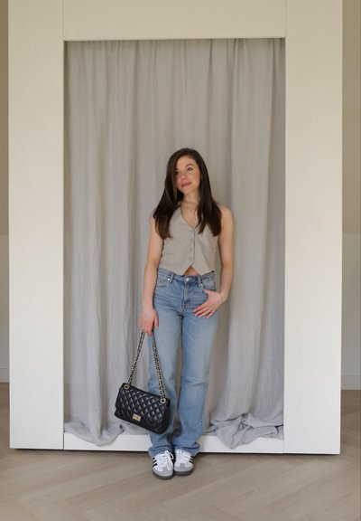 Chaleco gris corto con botones, jeans de mezclilla azul claro, zapatillas blancas y grises, y un bolso negro acolchado con cadena. Fondo suave gris.