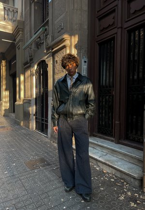 Chaqueta bomber de cuero negro sobre una camisa blanca y corbata estampada, combinada con pantalones de pierna ancha a rayas marino y zapatos negros, al aire libre.