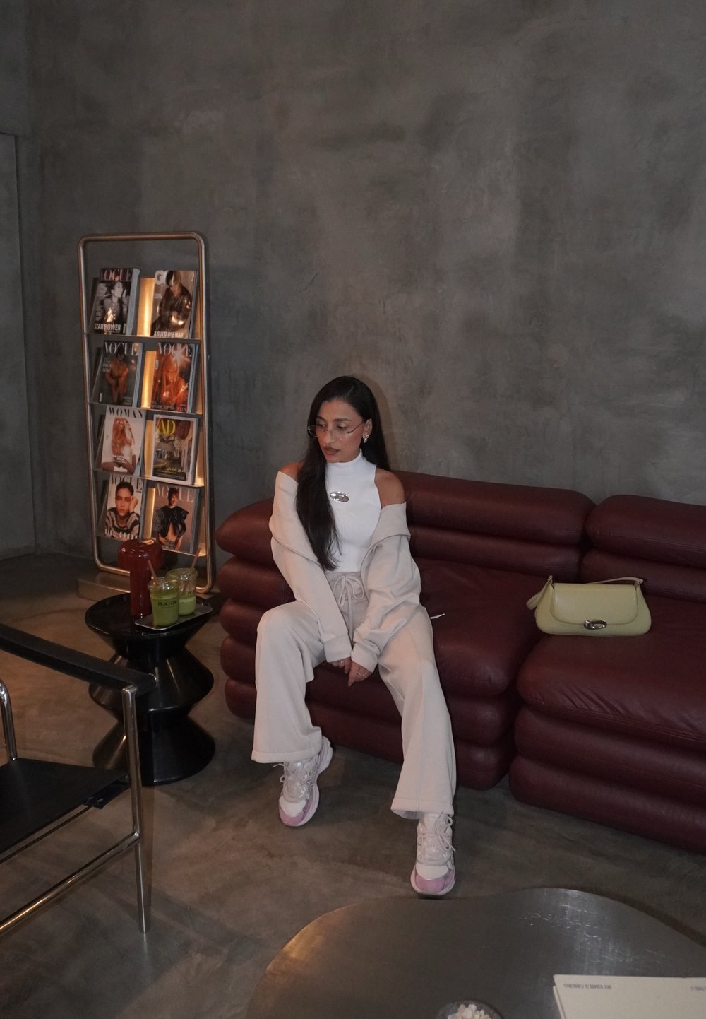 Femme en tenue blanche et baskets assise sur un canapé bordeaux dans une pièce minimaliste avec un porte-revues et une table portant des boissons.