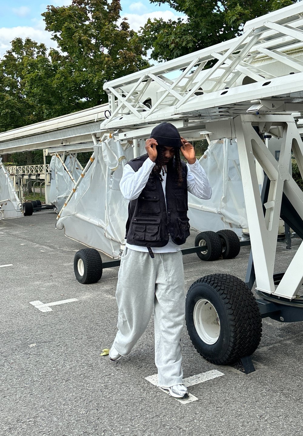 Μαύρο γιλέκο με πολλές τσέπες πάνω από ένα λευκό πουκάμισο, γκρι φόρμες και ένα μαύρο μπερέ, περπατώντας κοντά σε ένα λευκό όχημα με μεγάλα τροχούς.