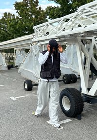 Μαύρο γιλέκο με πολλές τσέπες πάνω από ένα λευκό πουκάμισο, γκρι φόρμες και ένα μαύρο μπερέ, περπατώντας κοντά σε ένα λευκό όχημα με μεγάλα τροχούς.